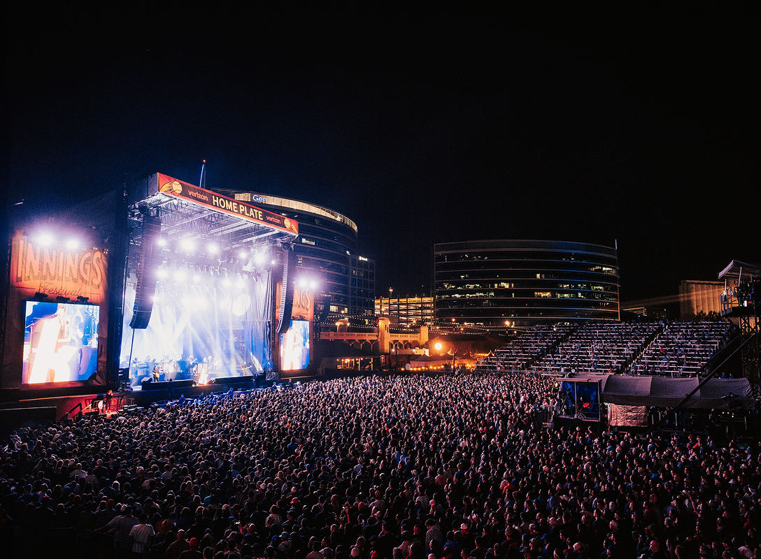 Large crowd watching a outdoor concert on a brightly lit stage.