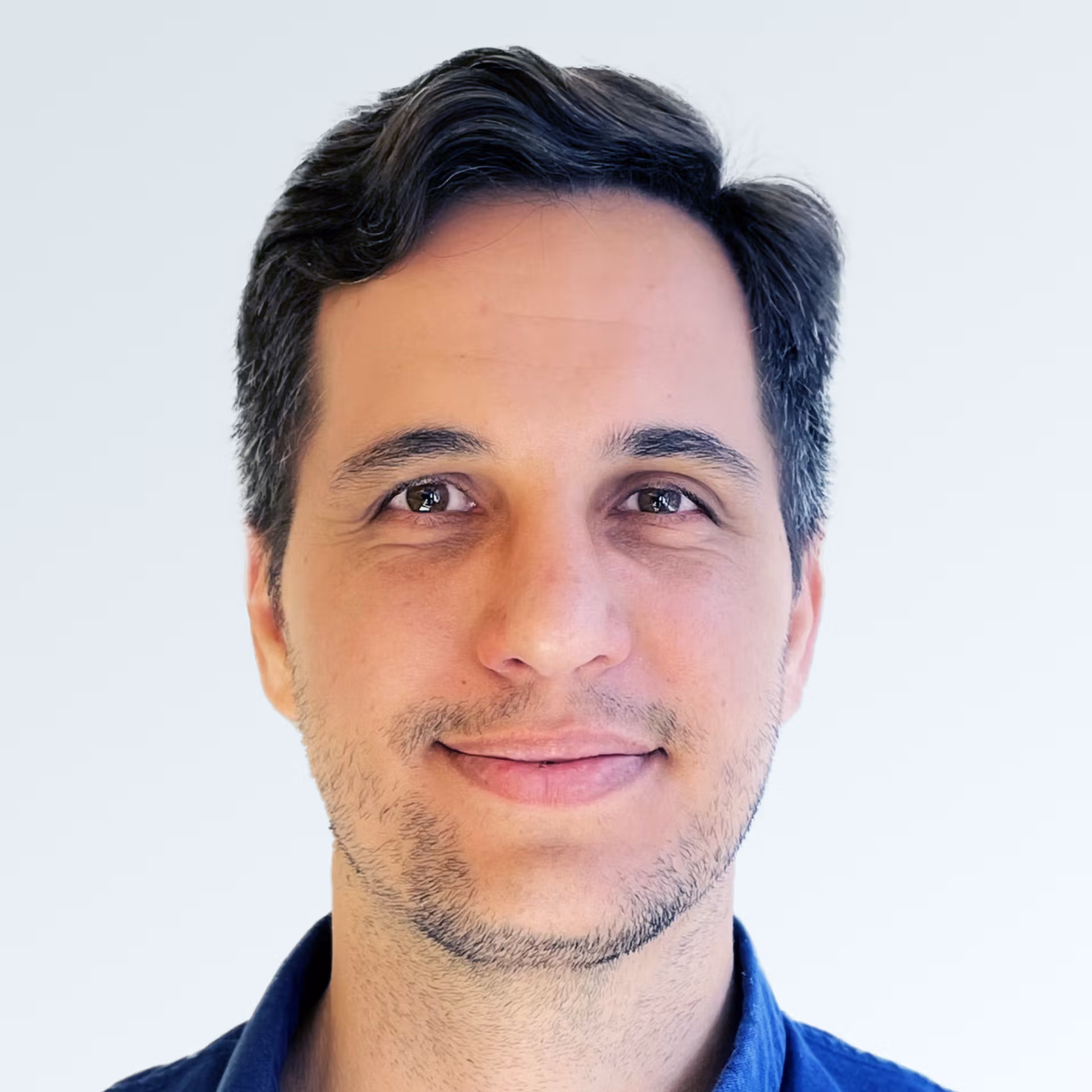Close-up portrait of a man with short dark hair and light stubble, smiling gently against a plain light background, wearing a dark blue shirt.