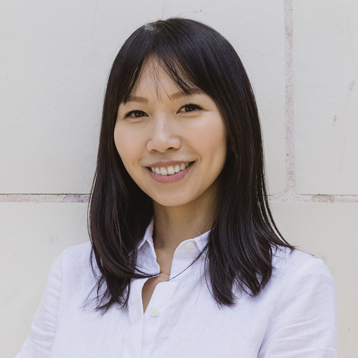 Smiling woman with straight black hair wearing a white button-up shirt standing against a light-colored tiled wall.