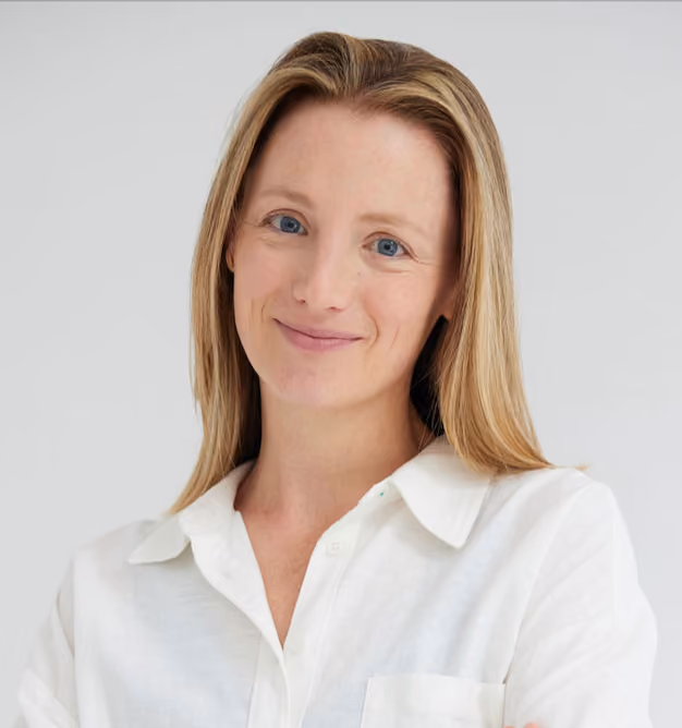 Smiling woman with straight blonde hair wearing a white collared shirt against a plain light background.