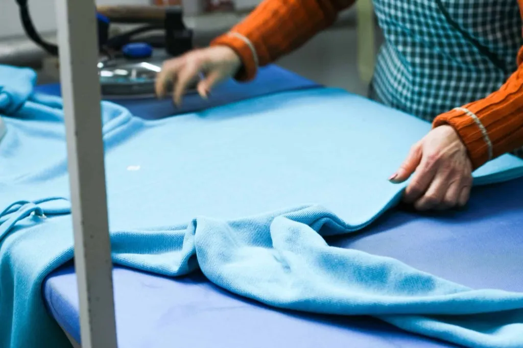 Hände streichen im Partnerbetrieb in Portugal ein türkisfarbenes Strickteil auf einem Finishtisch glatt, daneben ein Bügeleisen