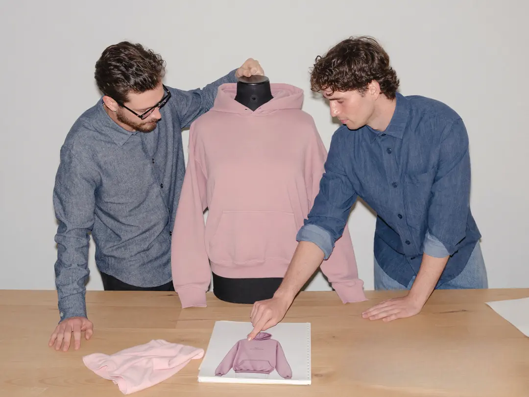 Zwei Männer prüfen einen rosa Hoodie an der Schneiderbüste und vergleichen ihn mit der Entwurfszeichnung auf dem Holztisch