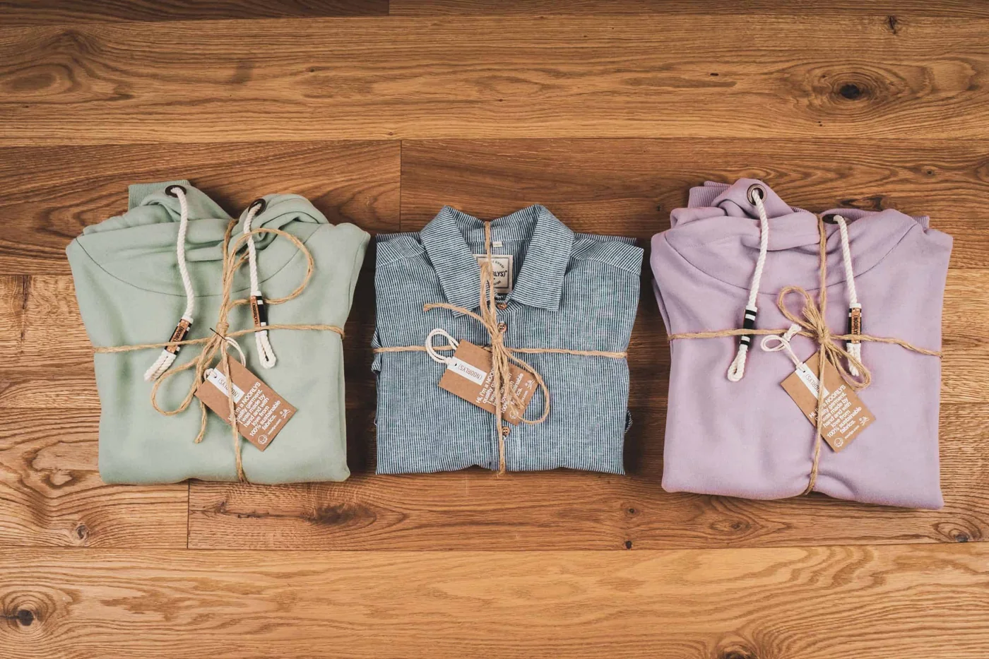 Zwei gefaltete Hoodies und ein gestreiftes Hemd mit Kordeln und Hangtags nebeneinander auf einem Holztisch.