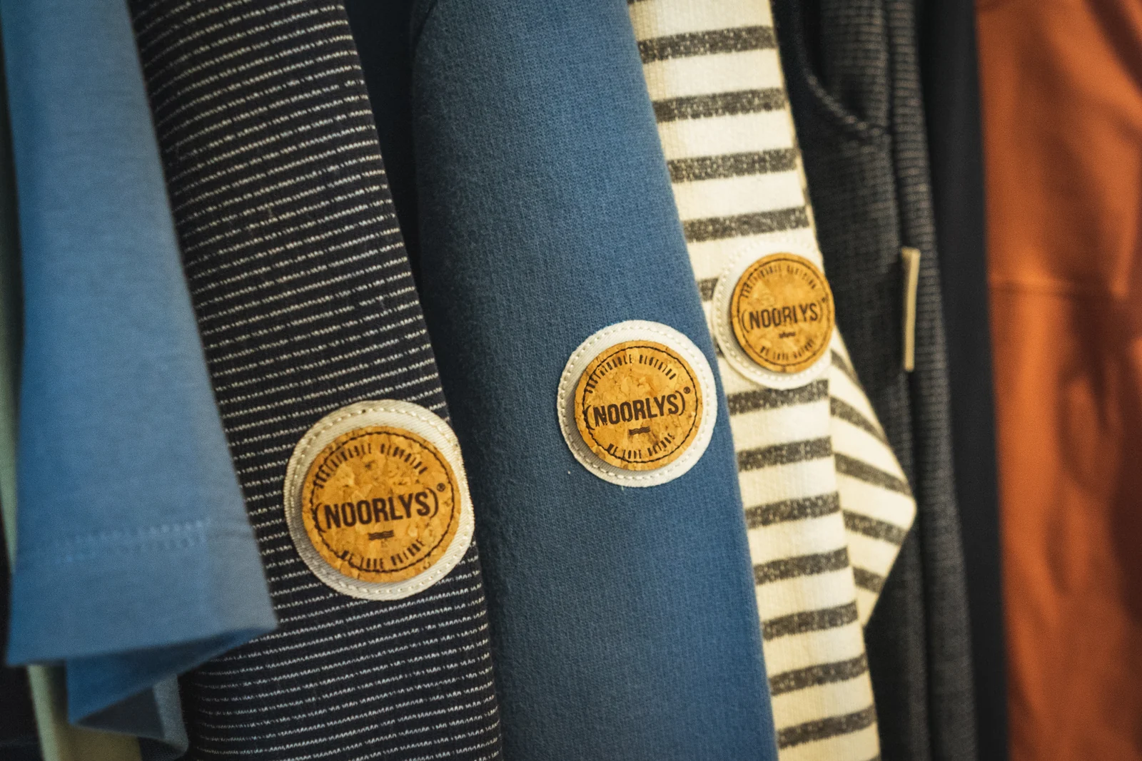Drei runde NOORLYS-Markenpatches auf blauen und gestreiften Textilien.