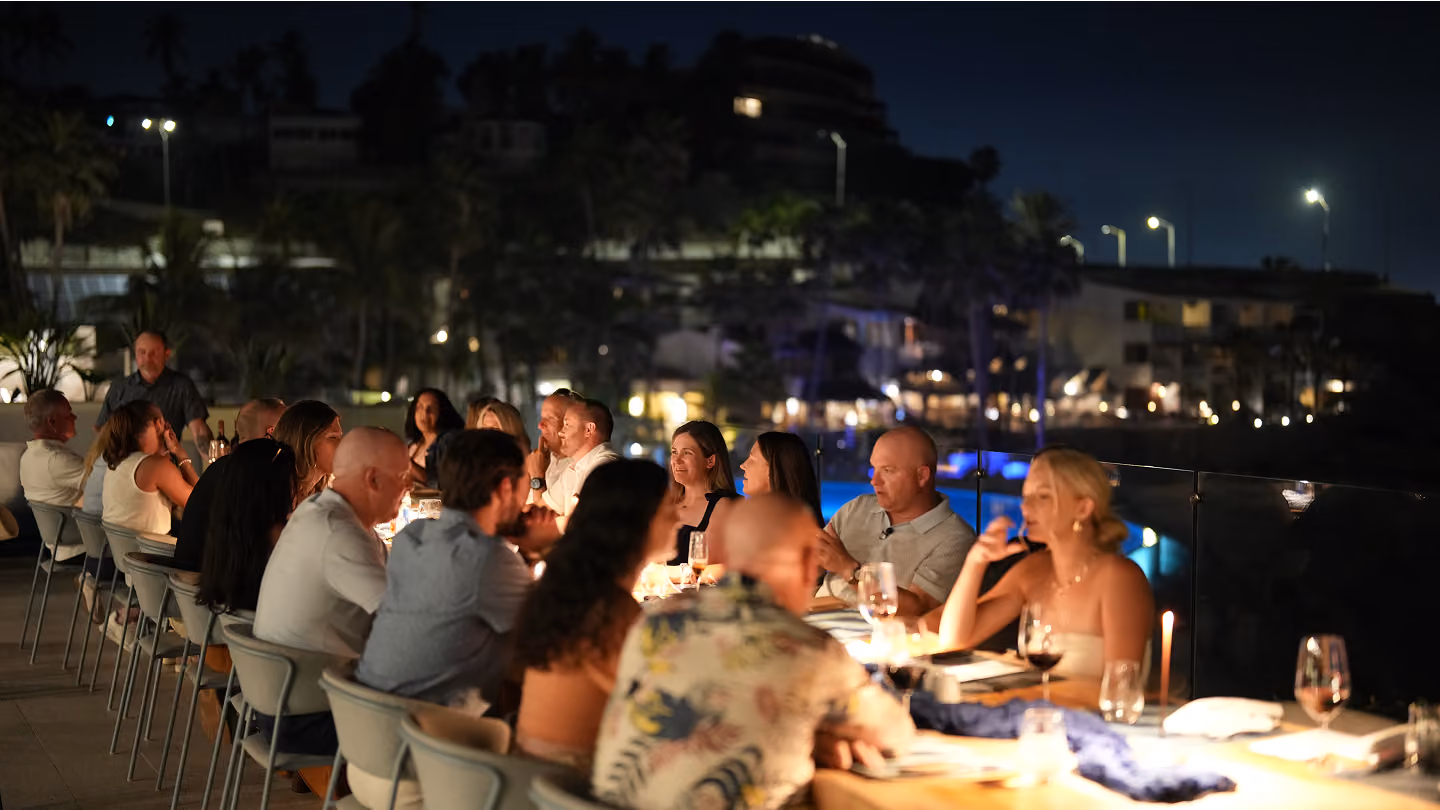 Group of people dining together outdoors at night with city lights and pool in the background.
