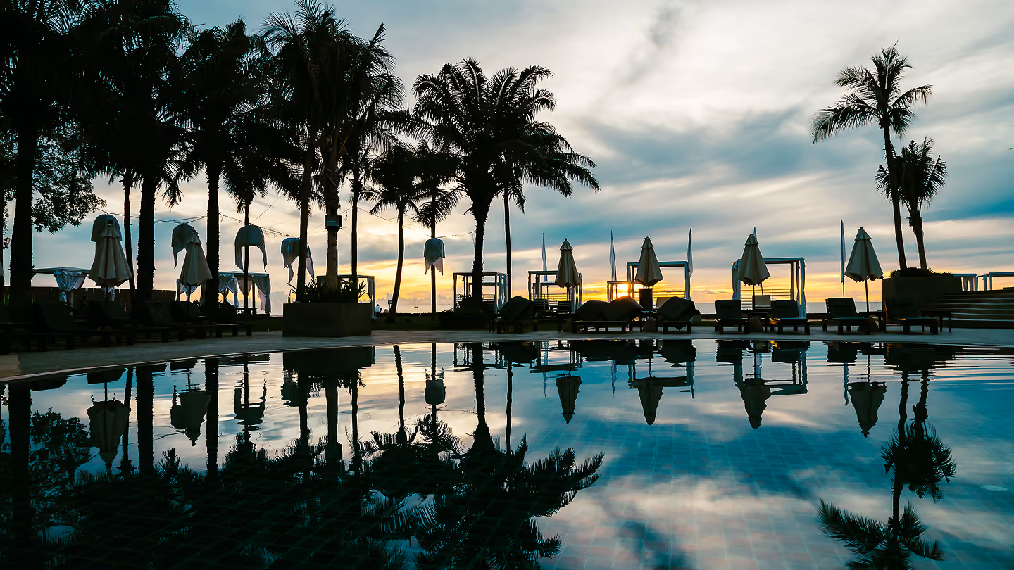 Palm trees and covered lounge chairs around pool reflecting sunset sky on a beach resort.