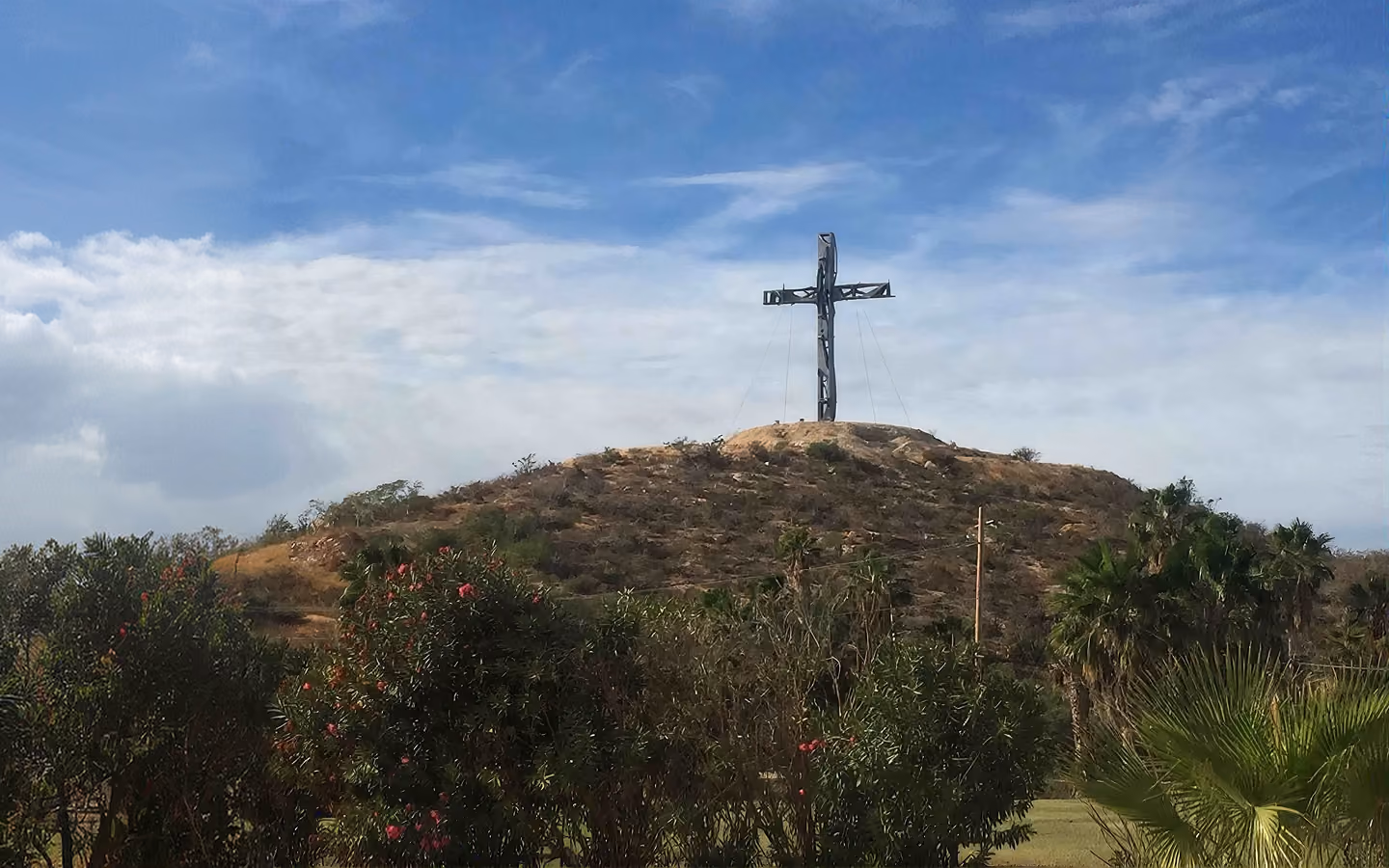 Large metal cross on top of a dry hill with bushes and palm trees under a partly cloudy sky.