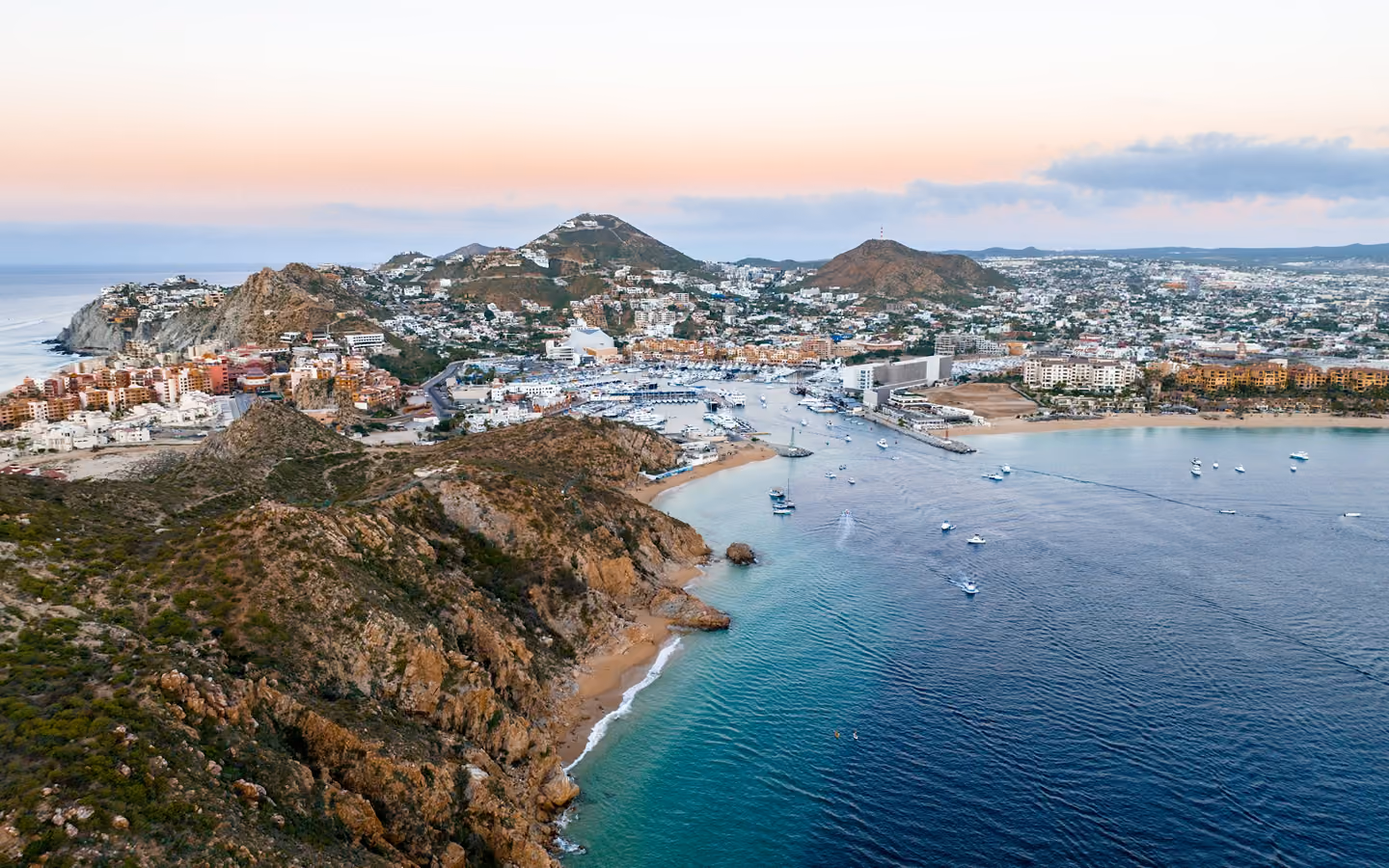 Aerial view of a coastal town with hills, buildings, marina, boats, and calm blue ocean water.