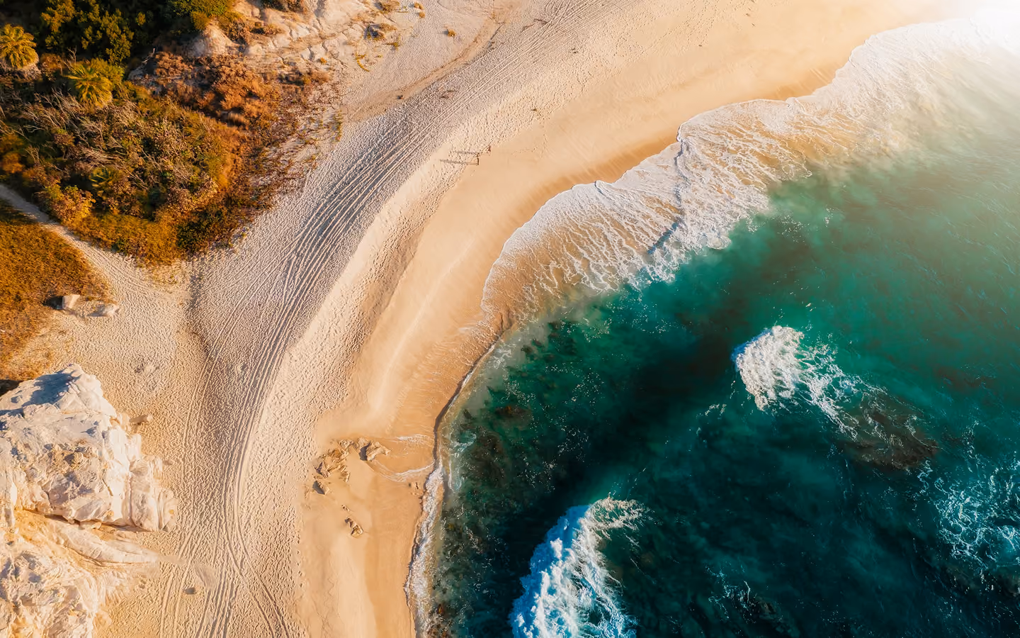 Aerial view of a sandy beach with waves from turquoise ocean washing ashore near rocky vegetation.