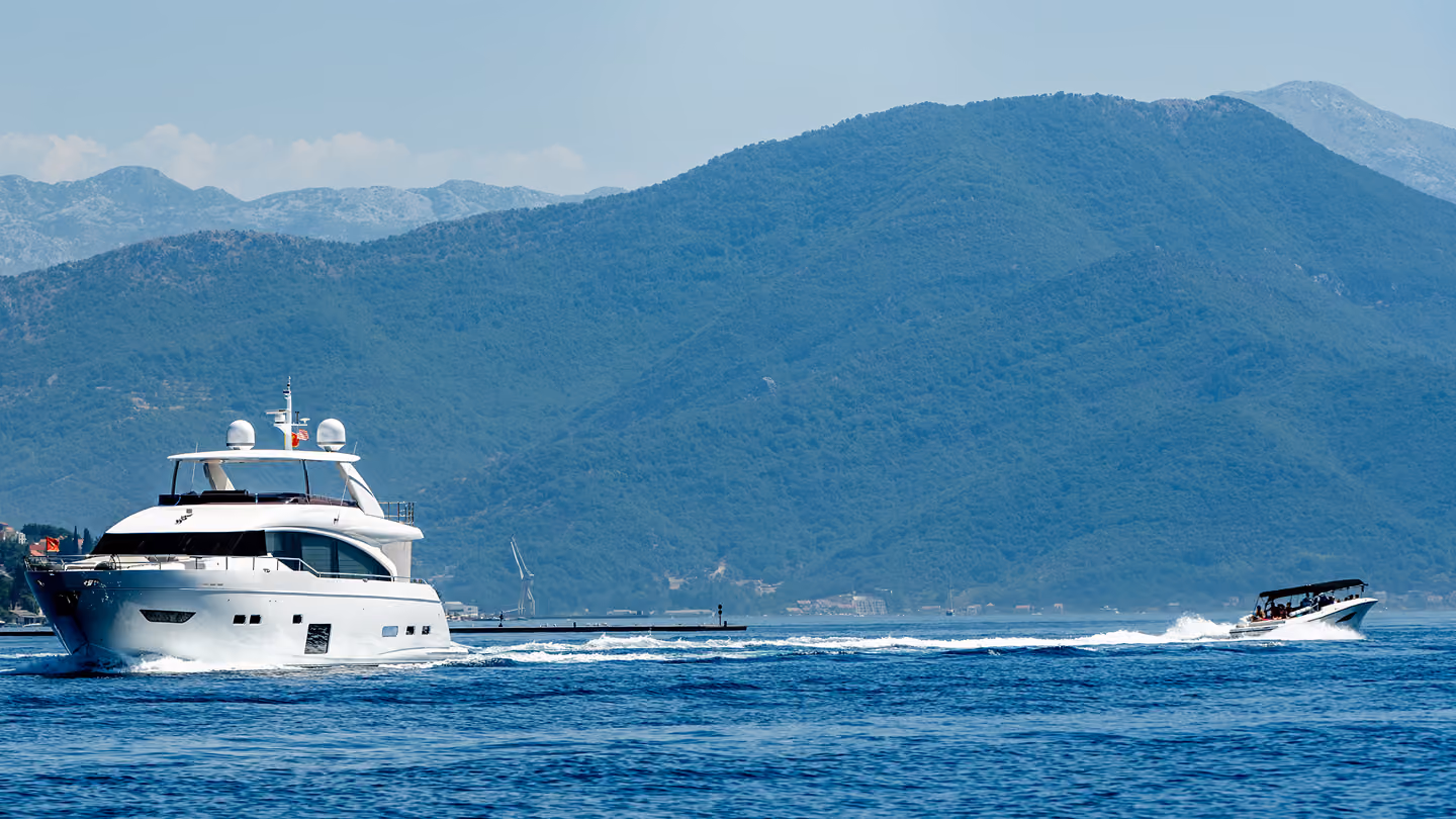 Two boats on blue water, one large yacht and one smaller speedboat, with green mountains in the background.