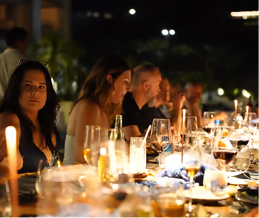People dining outdoors at night with candles and glasses on a long table.