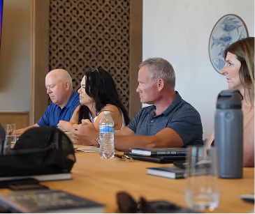 Four adults seated side by side at a table in a meeting room with notebooks and water bottles.