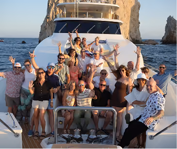 Group of people smiling and waving on a boat with rocky cliffs and ocean in the background.