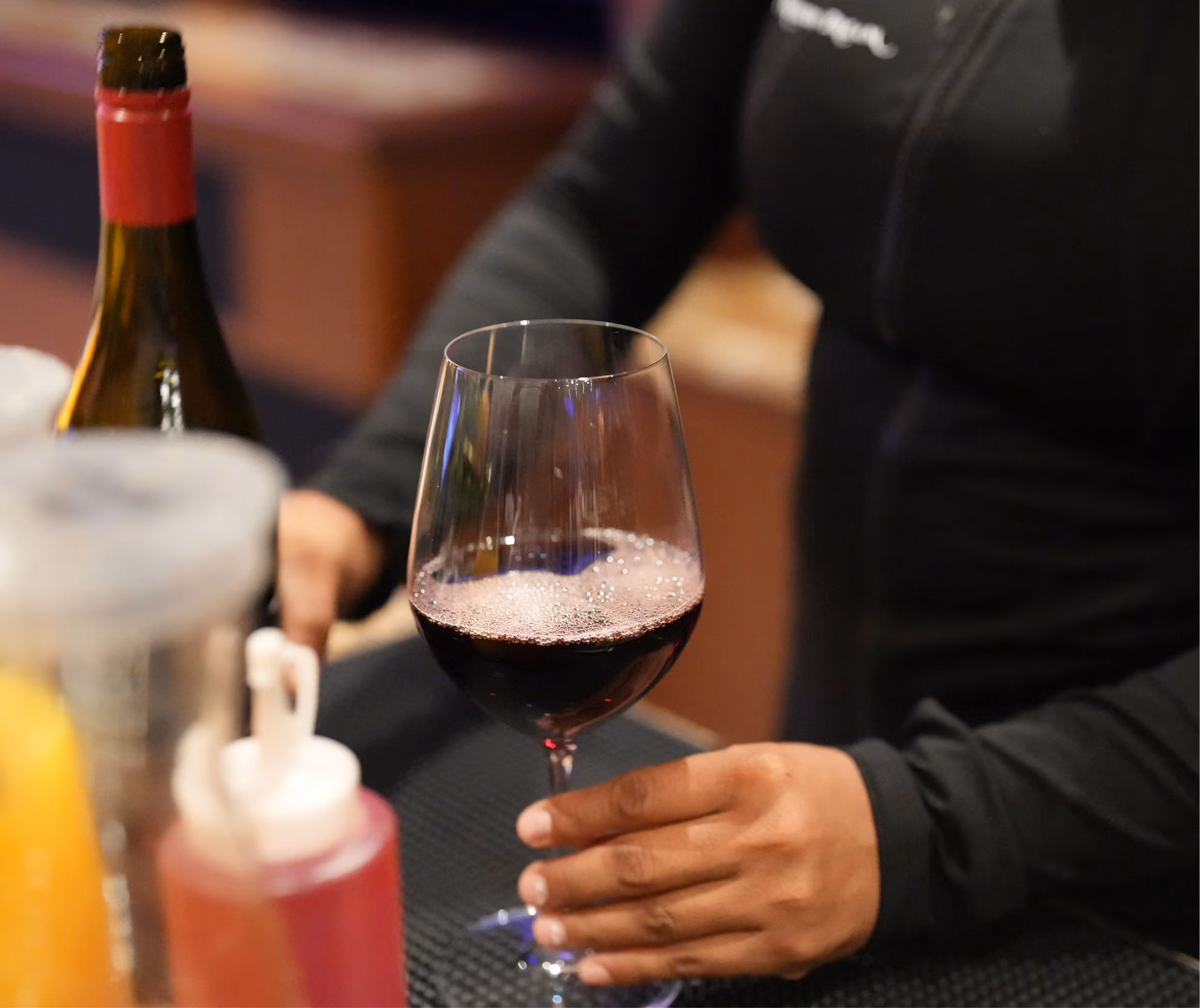 Person holding a glass of red wine at a bar with a wine bottle and condiments nearby.