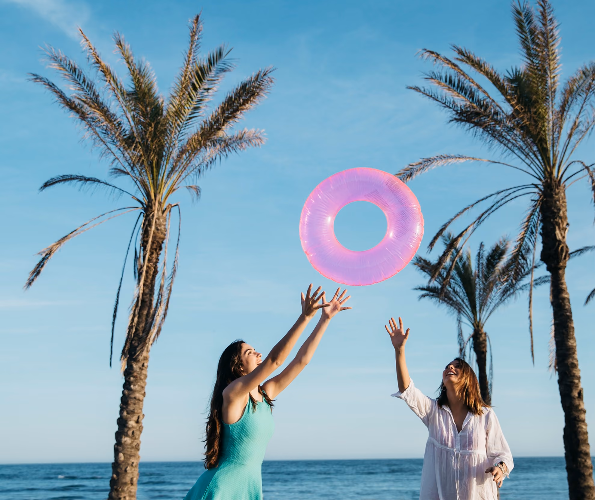Two women on a beach near palm trees reaching up to catch a bright pink inflatable ring against a blue sky.