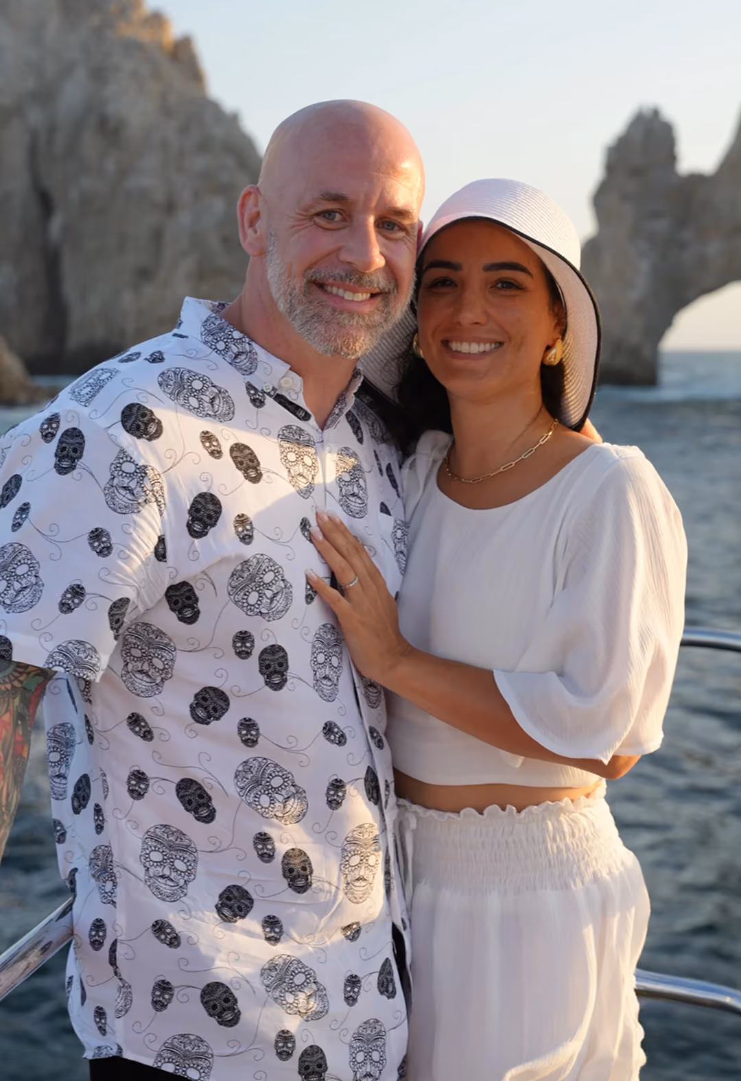 Smiling couple posing closely on a boat with ocean and rocky formations in the background during sunset; the man wears a white shirt with black skull patterns and has a beard and tattoos, the woman wears a white hat and a white outfit showing a ring on her left hand resting on the man's chest.
