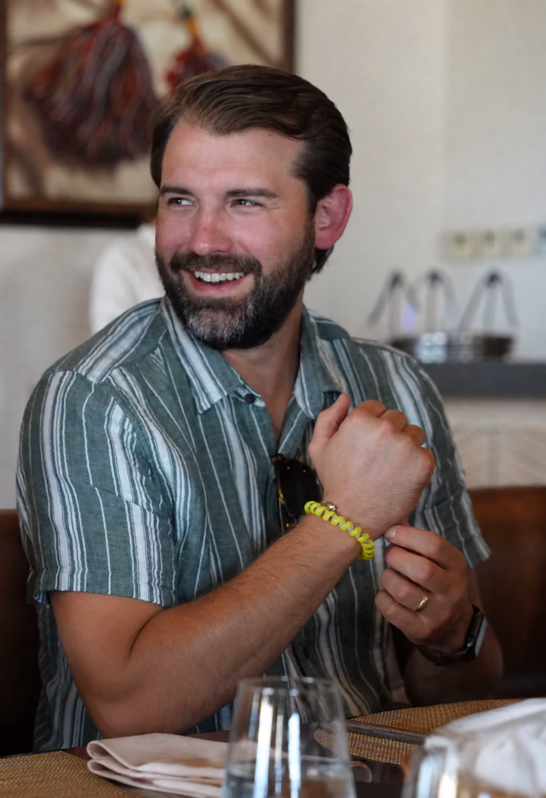 Man with beard and striped short-sleeve shirt smiling and adjusting a yellow spiral bracelet on his wrist while sitting at a dining table with a glass of water and napkin in the foreground.