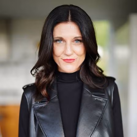 Portrait of a woman with medium-length dark hair and blue eyes, wearing a black turtleneck and sleeveless leather jacket, smiling slightly against a blurred indoor background.