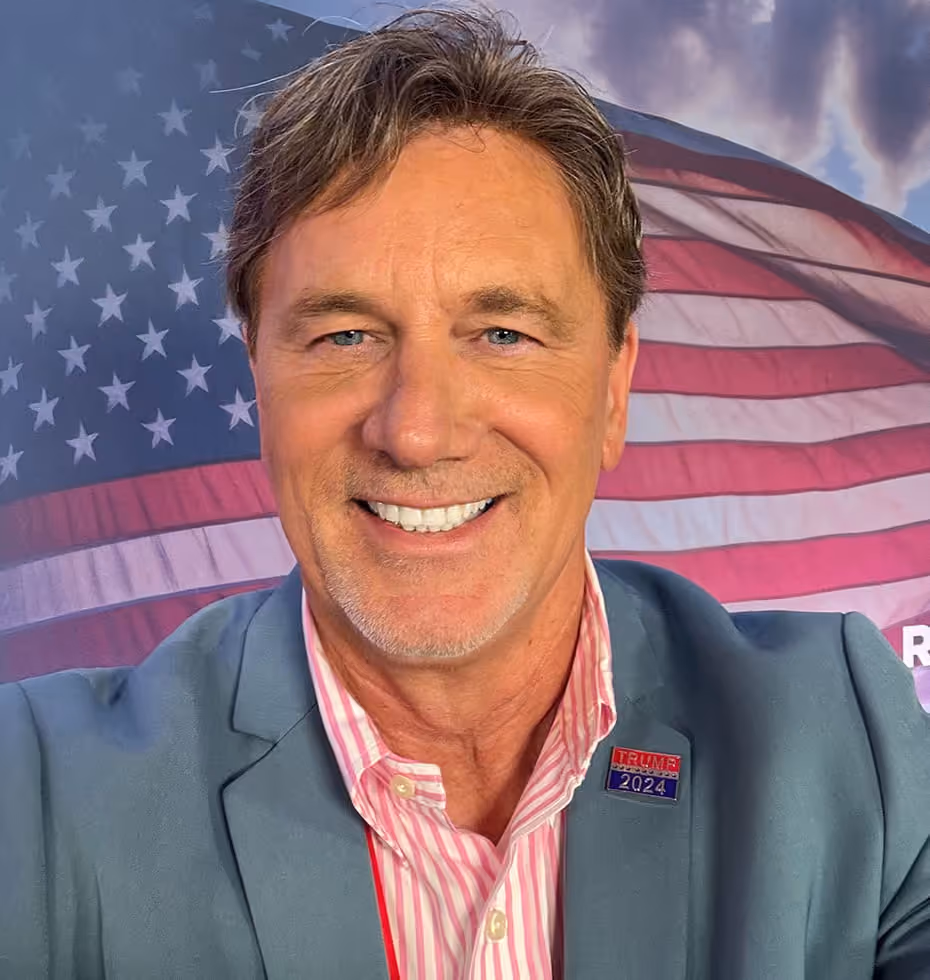 Smiling middle-aged man with light blue eyes and graying hair wearing a blue blazer and pink striped shirt, with a Trump 2024 pin on his lapel, in front of a waving American flag backdrop.
