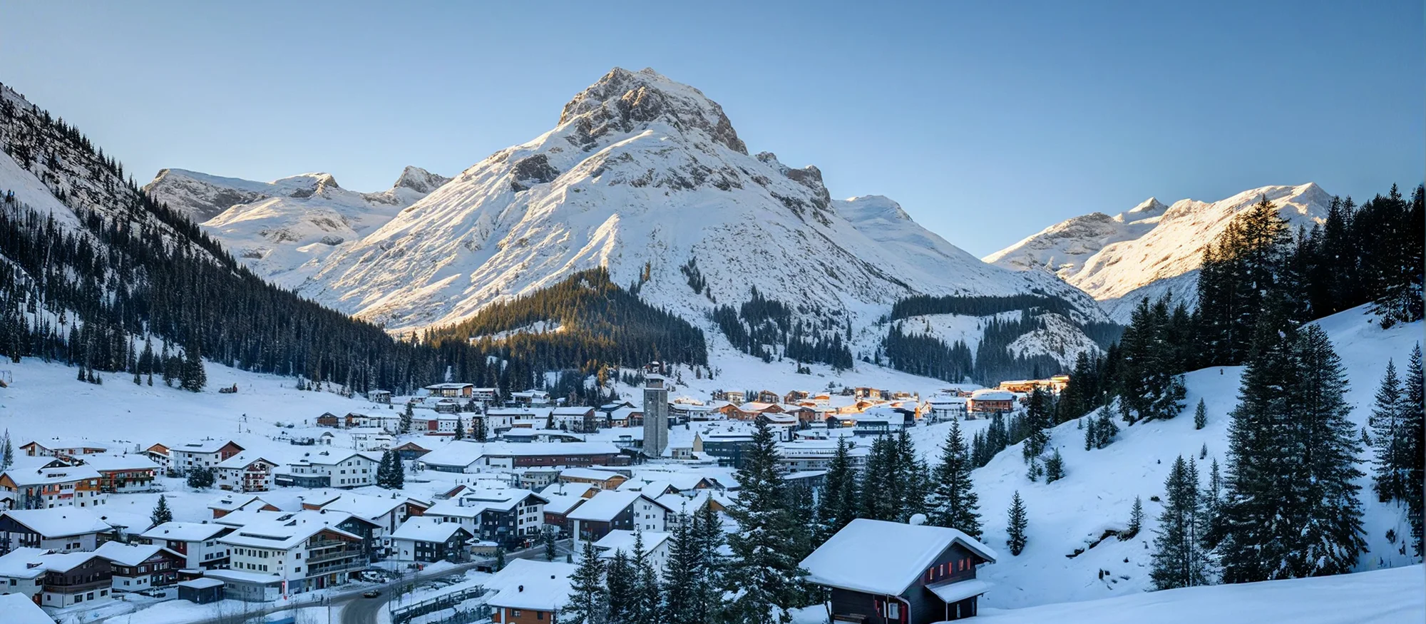 Snow-covered alpine village at the base of tall, sunlit snowy mountains under a clear blue sky.