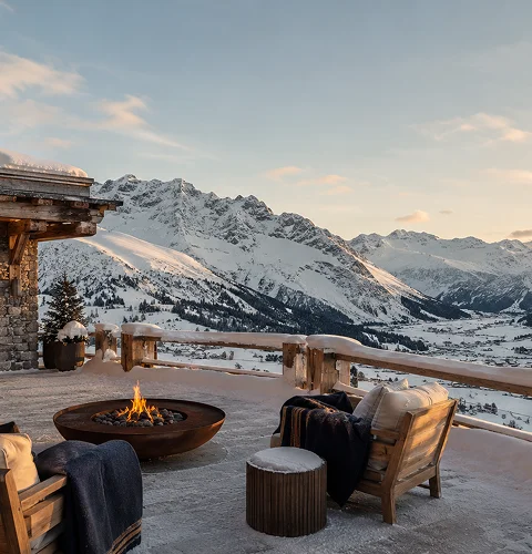 Mountain view balcony with fire pit, wooden chairs, and snowy peaks under a clear sky at sunset.