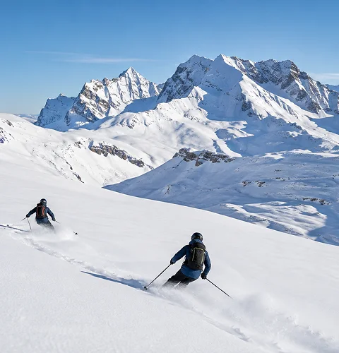 Two skiers descending a snowy mountain slope with snow-covered peaks under a clear blue sky.