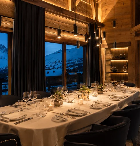 Elegant dining table set with glasses, candles, and flowers inside a wooden room with mountain view.