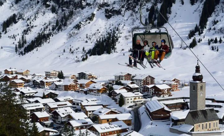 Three skiers riding a chairlift above a snowy alpine village with wooden houses and a church.