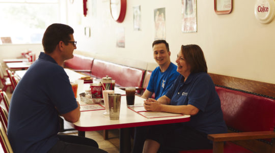 Three people in blue shirts sitting and chatting at a diner booth with drinks on the table.