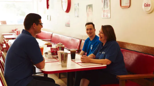 Three people in blue shirts sitting and chatting at a diner booth with drinks on the table.