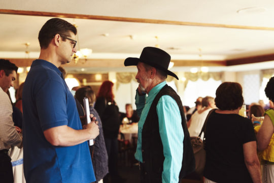 Two men engaged in conversation in a crowded indoor event space with people in the background.