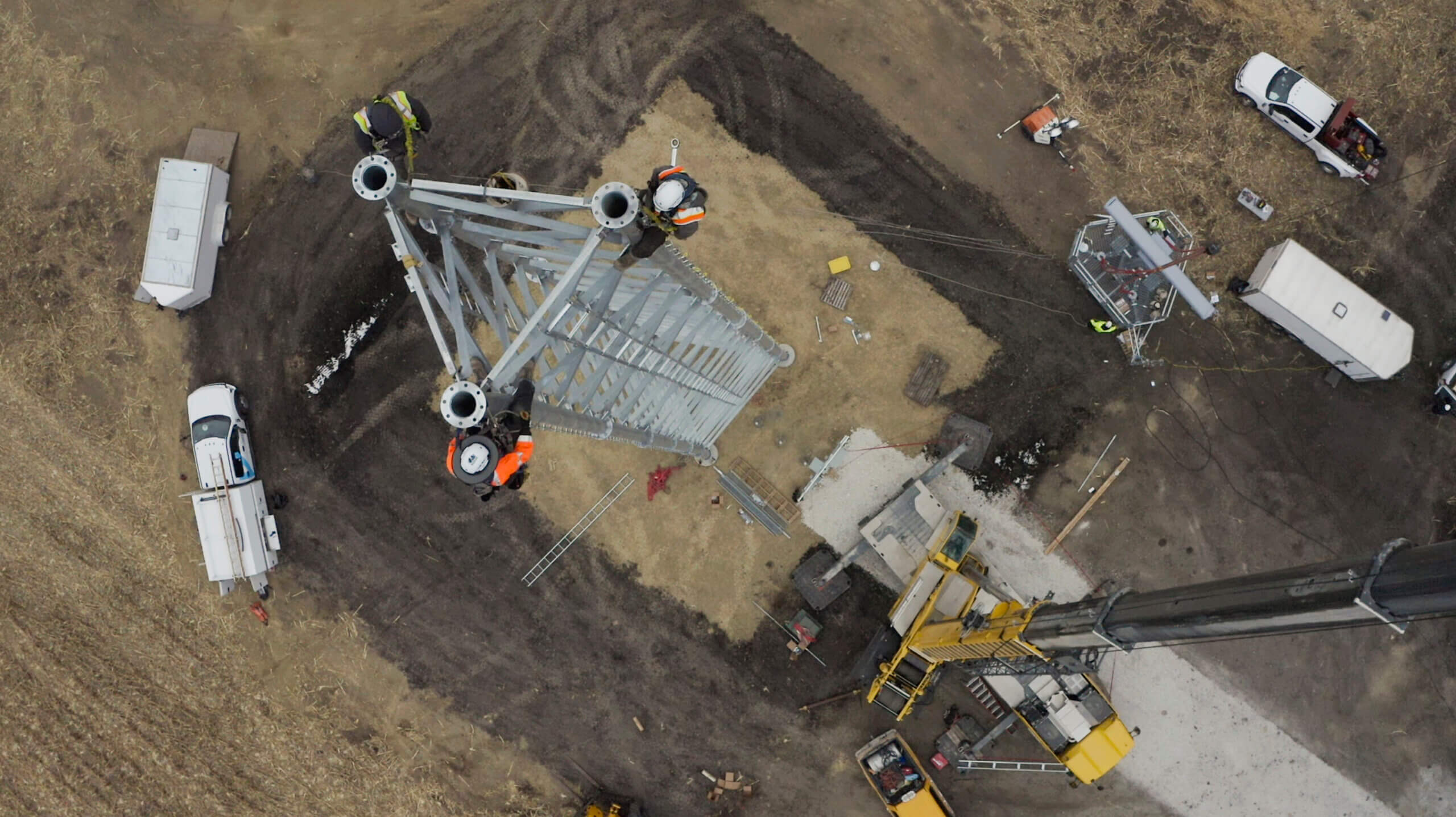 Construction workers assembling a tall metal tower viewed from above on a dirt site with vehicles nearby.