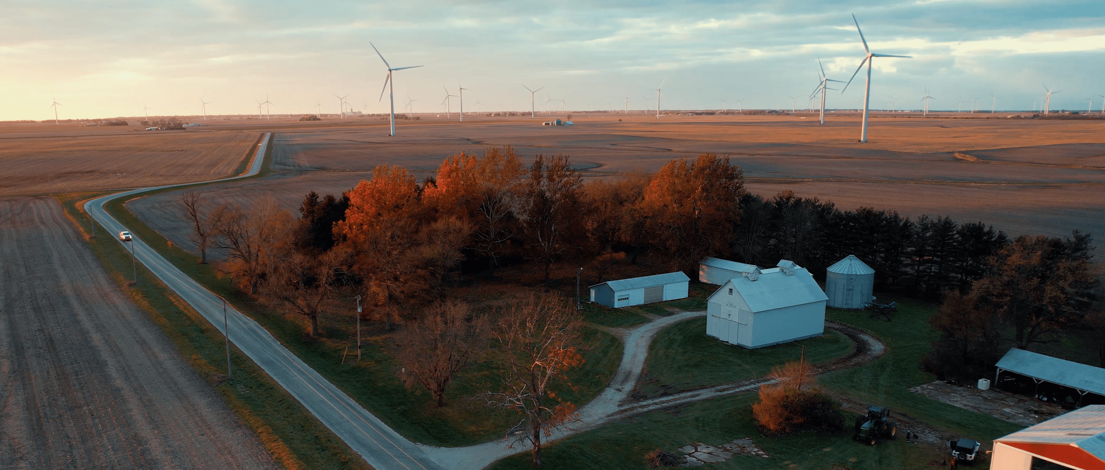 Rural farm with white barns, surrounded by trees and wind turbines in the distance at sunset.