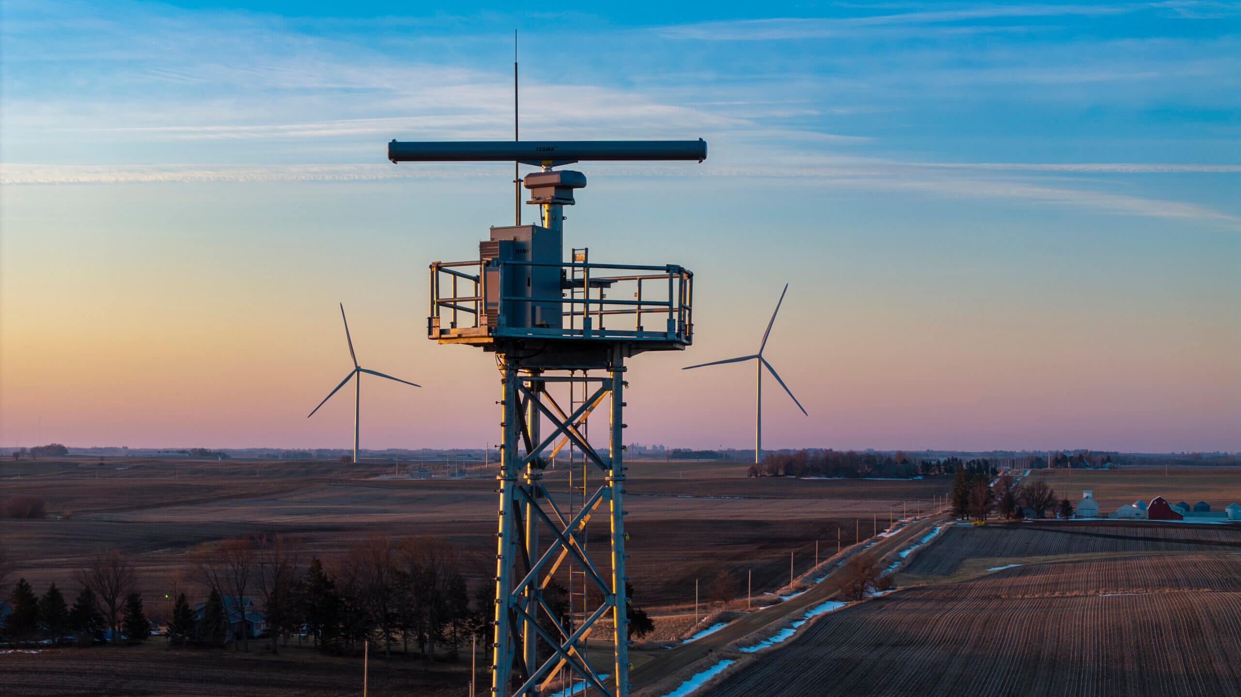 Metal tower with radar equipment in a rural area with wind turbines and farmland at sunset.