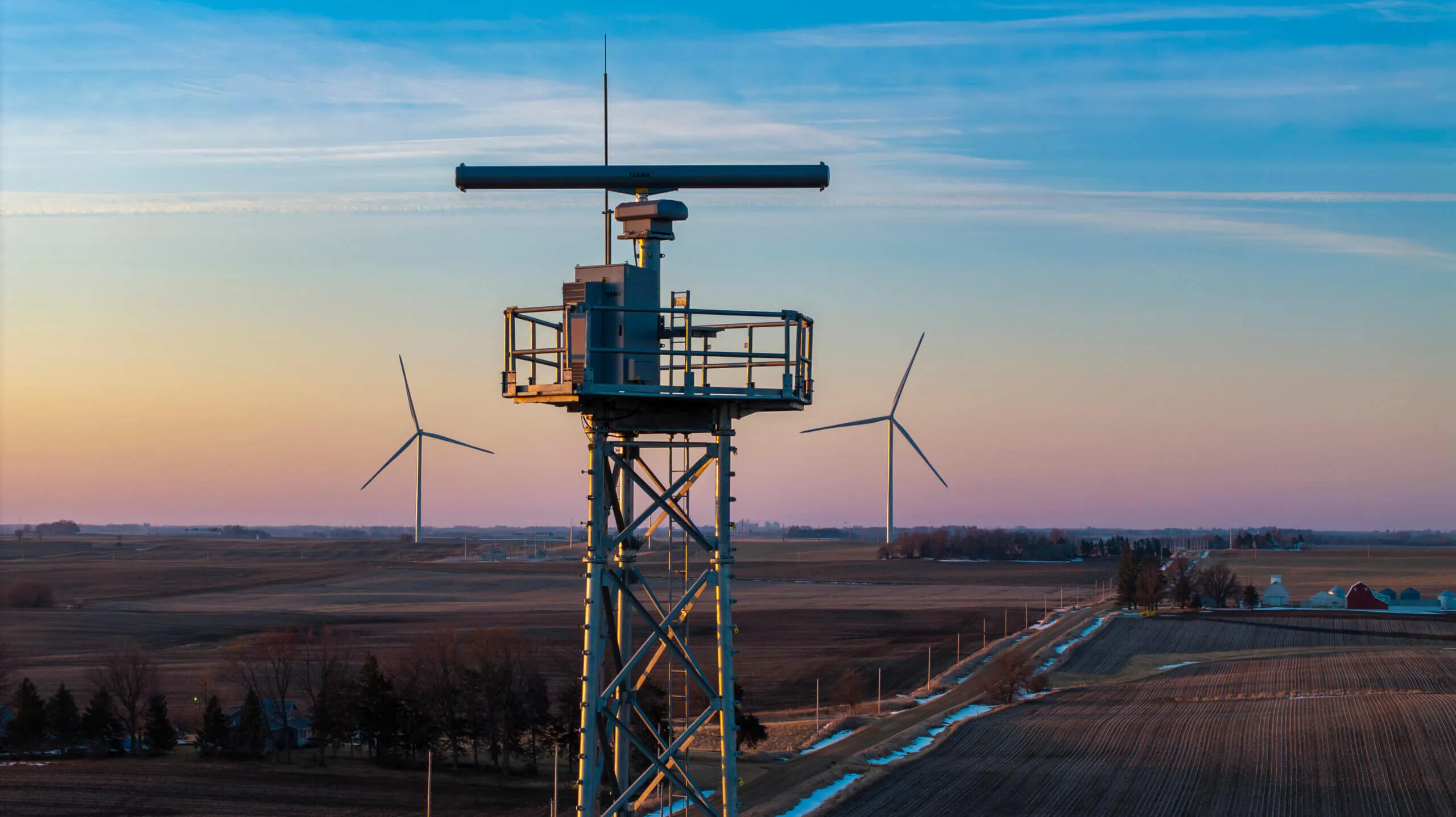 Metal tower with radar equipment in a rural area with wind turbines and farmland at sunset.
