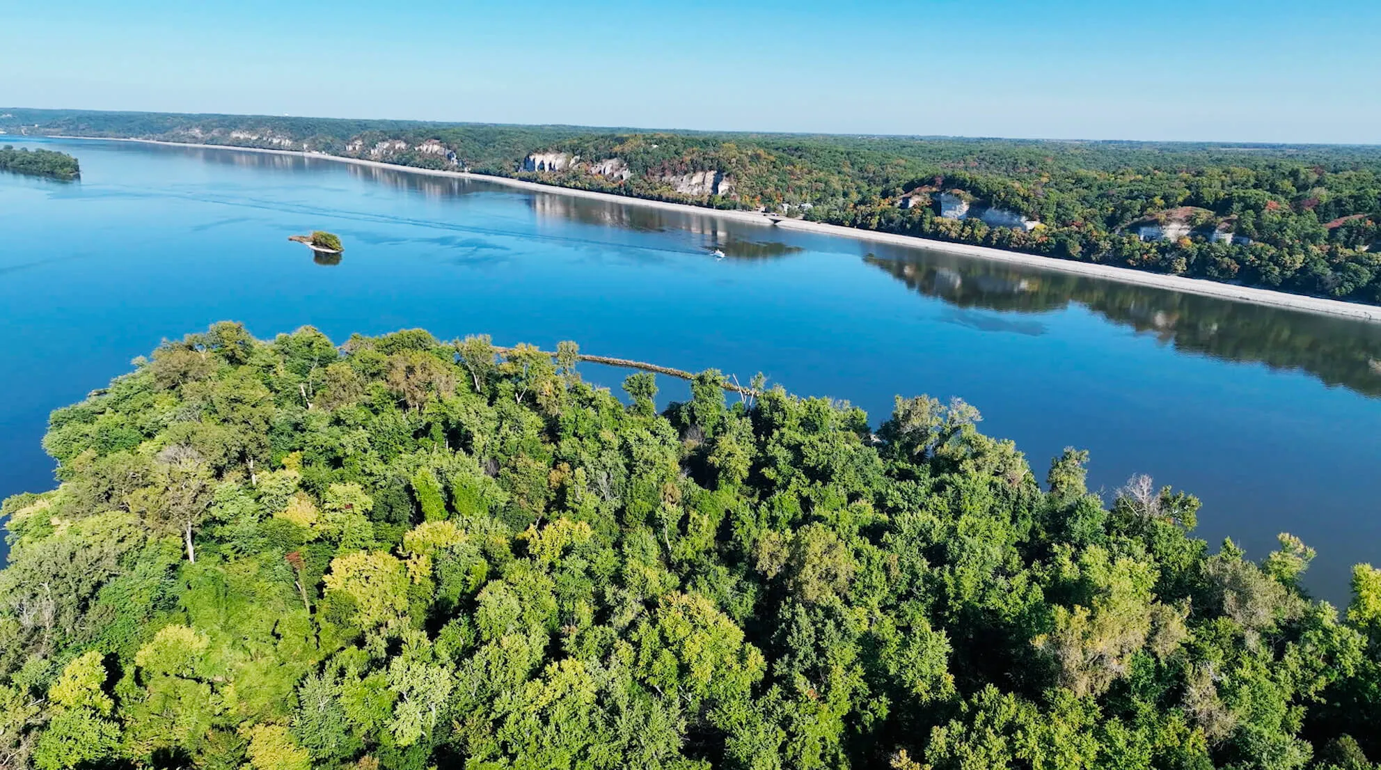 Aerial view of a lush green forested island bordered by a wide, calm blue river under a clear sky.