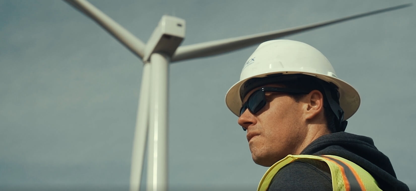 Man in a hardhat and safety vest with sunglasses framed by a large wind turbine.