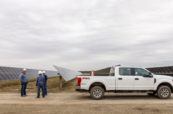 Three workers wearing helmets standing near solar panels and a white pickup truck on a dirt road.