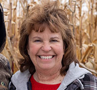 Smiling woman with brown hair wearing a red shirt and jacket stands in front of dry corn stalks.