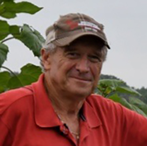 Smiling older man wearing a beige cap and red shirt outdoors near green plants.