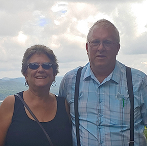 Two people outdoors with cloudy sky and mountains in the background, one wearing sunglasses and the other plaid shirt.