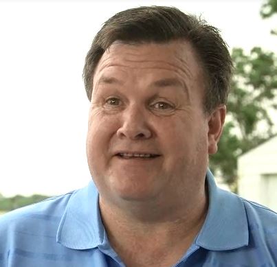 Smiling middle-aged man with brown hair wearing a light blue collared shirt outdoors.