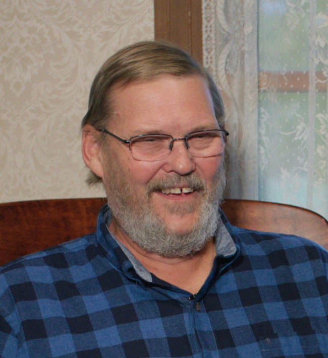 Smiling middle-aged man with glasses and a beard wearing a blue plaid shirt seated indoors.
