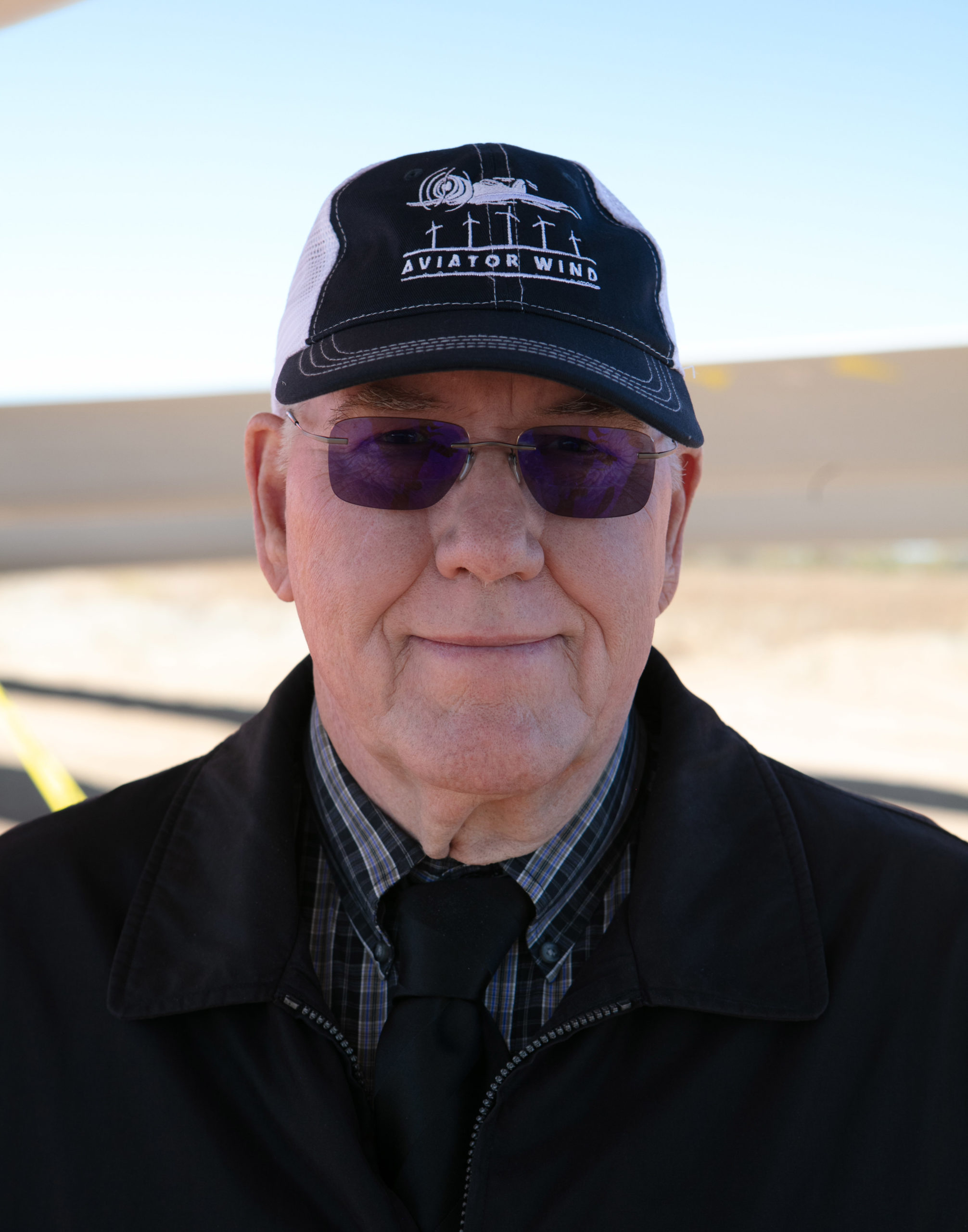 Older man wearing purple glasses, a black and white cap reading Aviator Wind, black jacket, and tie.