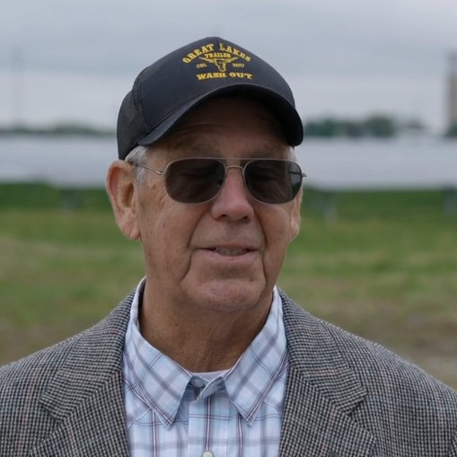 Older man wearing sunglasses, a cap, plaid shirt, and tweed jacket outdoors with blurred background.