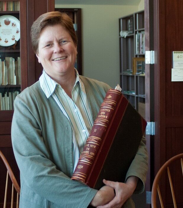 Smiling woman in a green jacket holding a large red and black book in a library setting.