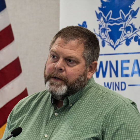 Man with beard wearing green checked shirt speaking in front of flag and blue logo on wall.