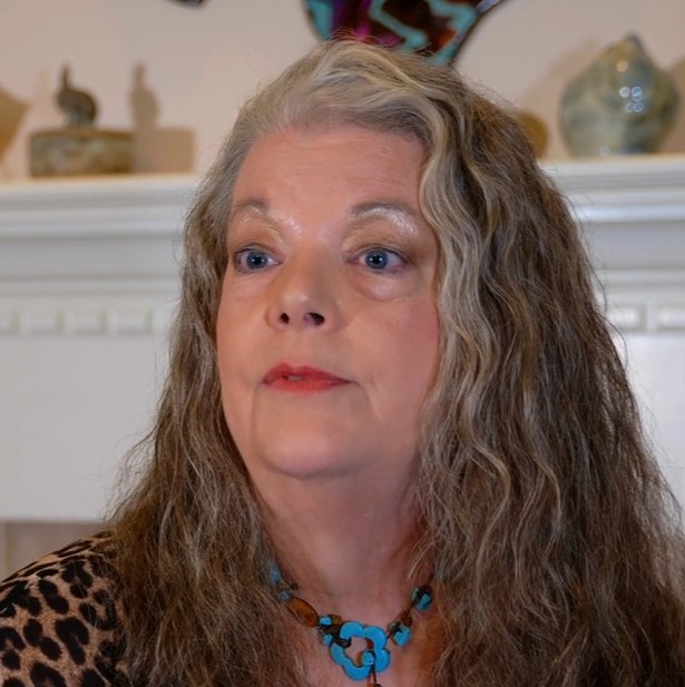 Older woman with long wavy gray hair and blue eyes, wearing a leopard print top and turquoise necklace.