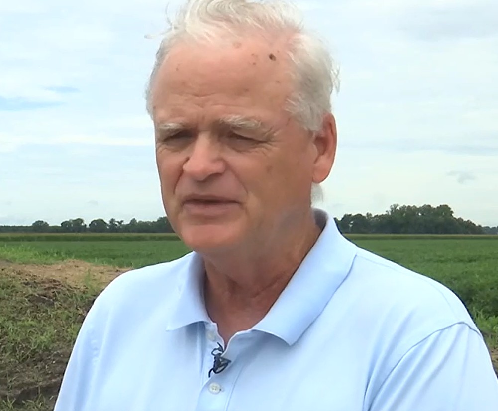 Older man with white hair in a light blue polo shirt standing outside with green fields behind him.