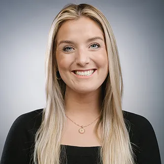 Smiling woman with long blonde hair wearing black top and gold necklace against a gray background.